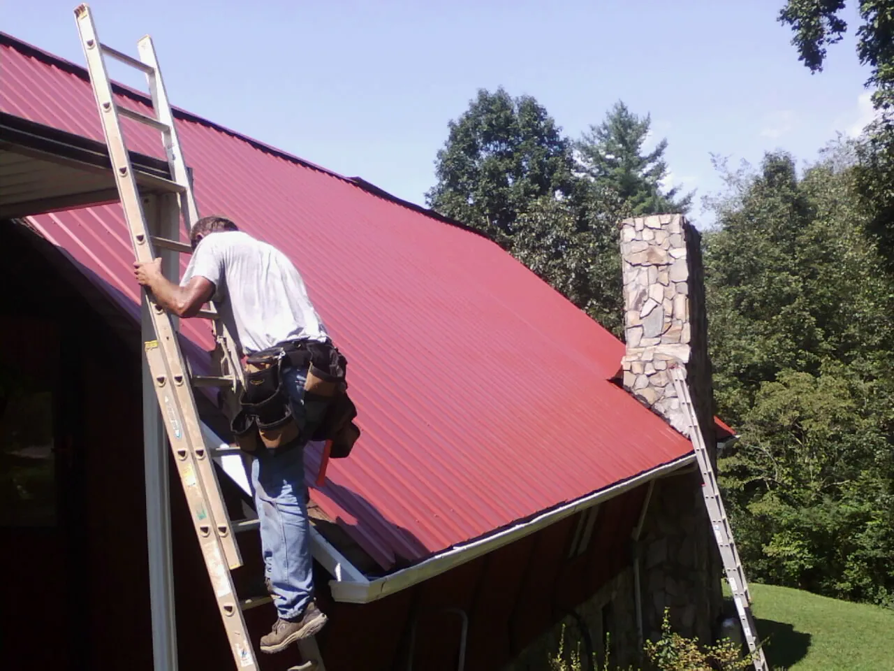 Roofer on a ladder beside a red metal roof and stone chimney — Soffit & Fascia Repair by Roofing Brothers in Berwick